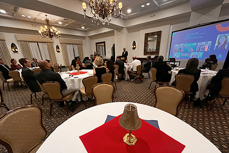 [A table with red-blue squares with candle in foreground and people seated at tables listening to a South Asian man speaking at a podium and watching a screen shot of Tara Carr, MD, with photos from her life and career]