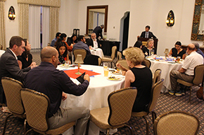 [Asian man in suit-and-tie at podium in front of tables of people eating as he begins to speak.]