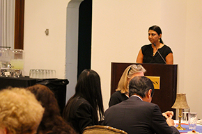 [Woman with dark hair and dark top at podium speaks as people eat at tables in foreground.]