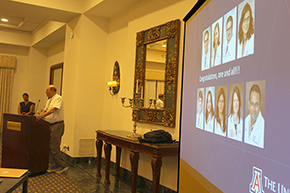 [Balding man in white shirt with bolo tie and khaki pants at podium and woman watching behind him with blue screenshot of photos of doctors promoted and the phrase, “Congratulations, one and all!!!” in foreground on right]