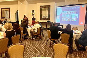 [Woman wearing maroon top at podium with another woman wearing dark top and red-and-black skirt behind to her left and, on the screen, the name and photos of Mohanad Al-Obaidi, MD, MBA, an infectious diseases physician in the U of A Department of Medicine]