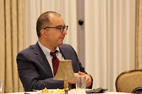 [Man with glasses in suit-and-tie sits at table watching as he’s being complimented on his hard work and dedication to the job.]