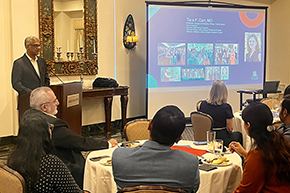 [South Asian man with gray hair and glasses in dark suit at podium on left with round tables of people in foreground and, above right, a screen with a blue slide up with the name and photos of Tara F. Carr, MD]