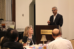 [South Asian man with gray hair and glasses in dark suit at podium with people in front of him at round tables.]