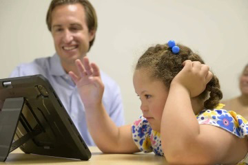 [Dan Combs is smiling in the background while a young girl looks at a computer screen.]