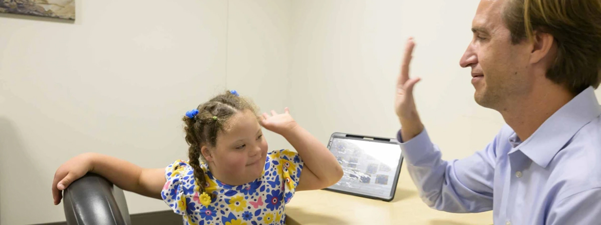 [Dan Combs, MD, assistant professor in pediatrics and sleep medicine at the University of Arizona College of Medicine – Tucson’s Department of Medicine, holds his hand up to a young Down syndrome patient for a high five in an office.]