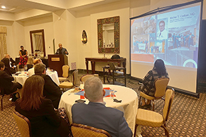 [Man with blue jacket gesturing at podium with woman in red-and-black skirt behind him and two round tables in front – with a screen showing name and photos of Michel T. Corban, MD]