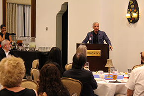 [Man with blue jacket at podium that says “Arizona Inn” with woman behind him to left and people sitting facing him from round tables in front of him]