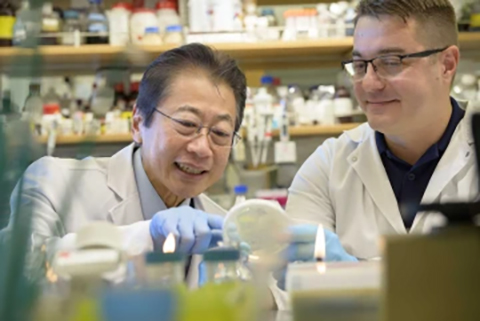 [Two men in white lab coats and glasses closely examine a petri dish while surrounded by bottles of various sizes.]