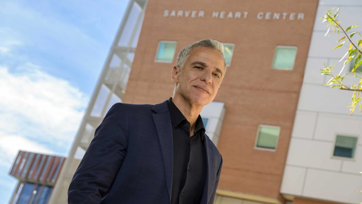 [A man in a dark suit coat and black shirt stands outside a building that has Sarver Heart Center signage.]