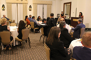 [Woman at podium on right talking to audience seated at round tables in large room with wall lamp sconces and beige drapes at the Arizona Inn]