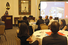 [Woman at podium on left talking to audience seated at round tables in large room with wall lamp sconces and a screen picturing name and photos of Sima Ehsani, MD]