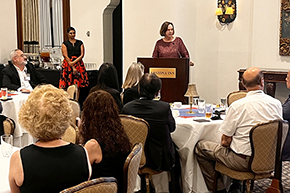 [Woman in maroon top at podium that says “Arizona Inn,” with another woman behind her in red-and-black skirt and people seated at round tables in front of her.]