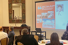 [People seated at tables around a screen with a red-and-white slide with name of “Justin F. Hayes, MD” and photos of him and his family and colleagues in the Division of Infectious Diseases.]