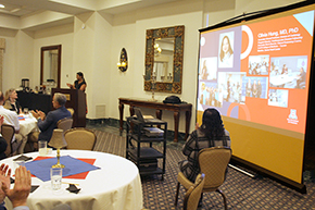 [Woman with dark top and red-and-black skirt at podium with people seated at round tables in front of her and, to her left, a woman seated next to a projector showing a slide on a screen that’s red and blue with name and photos of “Olivia Hung, MD, PhD”]