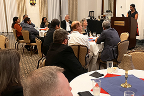 [Woman with dark top and red-and-black skirt at podium with people seated at round tables in front of her]