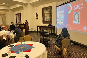 [Woman at podium on left talking to audience seated at round tables in large room with wall lamp sconces and a screen picturing name and photos of “Neha Jaswal, MD”]