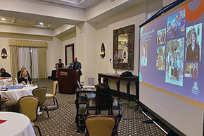 [Man with blue jacket at podium that says “Arizona Inn” with woman behind him to left, people sitting facing him from round tables, and a blue screen display at right with name and photos of “Elizabeth Juneman, MD”]