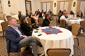 [Man and woman sit at table, facing to the right, with others listening to speaker talking from off-stage right]