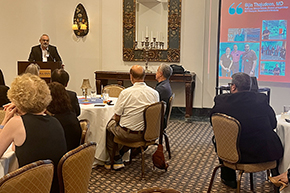 [A man with gray hair and glasses wearing a blazer talks to audience as screen displays name and photos of “Bijin Thajudeen, MD,” a professor in the Division of Nephrology at the U of A Department of Medicine.]