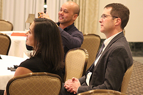 [Two men and a woman seated at a table listen to a presenter talk about faculty members advancement opportunities in the Department of Medicine at the U of A College of Medicine – Tucson.]