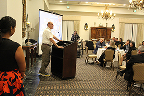 [Man in white shirt, bolo tie and khaki pants speaks from podium with people seated at round tables to his front and woman over his shoulder from behind wearing black top and red-and-black skirt as slide show on screen shows photos from prior year’s U of A Department of Medicine faculty promotion event.]