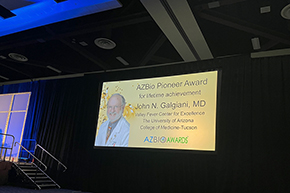 [A yellow illuminated slide is projected on a screen at the AZBio Awards Ceremony with the face of an older white man wearing a physician's white coat and emblazoned with the words: "AZBio Pioneer Award for lifetime achievement, John N. Galgiani, MD, Valley Fever Center for Excellence, University of Arizona.”]