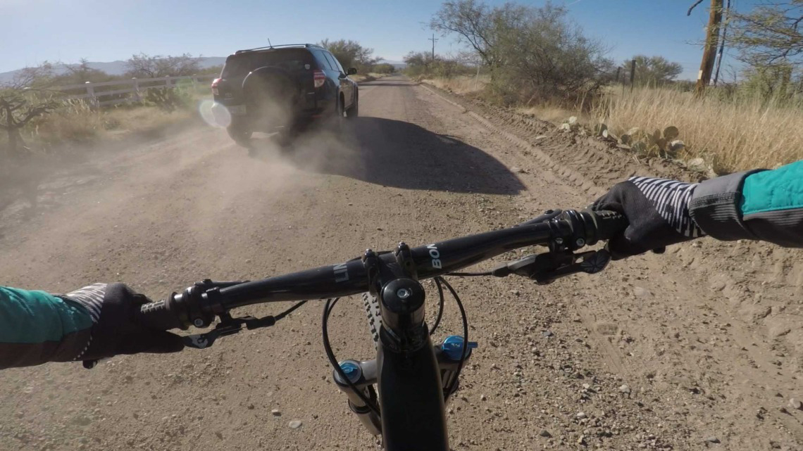 [A vehicle kicks up dust as it passes a bicyclist on a dirt road]