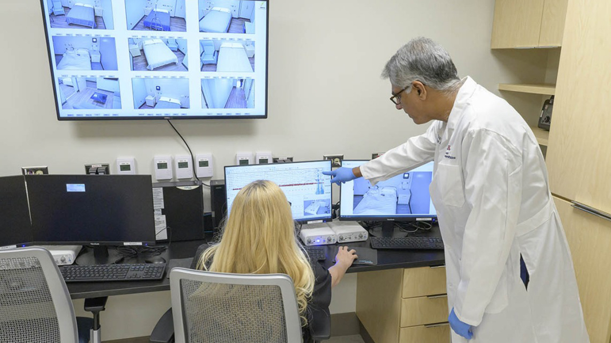 [Man wearing white coat stands pointing at a screen. A woman with long blonde hair sits at a desk in front of the computer screen the man is pointing at.]