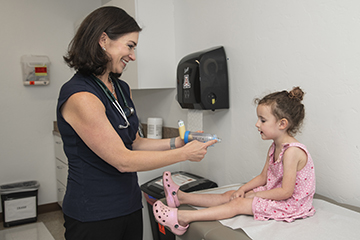 [A woman in dark dress wearing stethoscope shows a child sitting on an exam room table the nipple of a bottle]