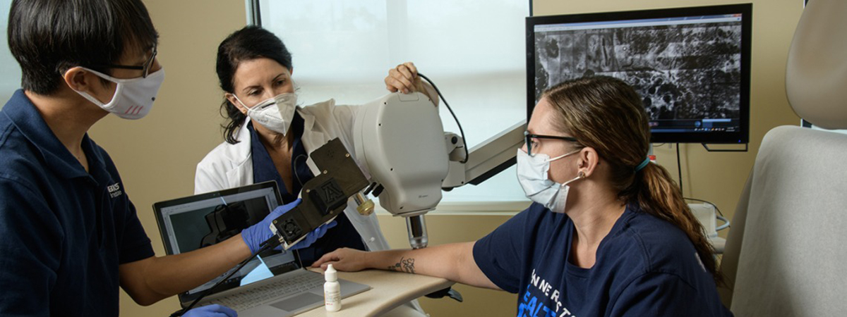 [Clara Curiel Lewandrowski, MD, and Dongkyun Kang, PhD, prepare a side-by-side test of their confocal microscope and a portable imaging unit to compare image quality. Dr. Curiel aligns the larger device, while Dr. Kang prepares to aim the handheld one at a nearby spot on Savannah Barber’s arm. Barber is a medical assistant at Banner – University Medicine Tucson.]