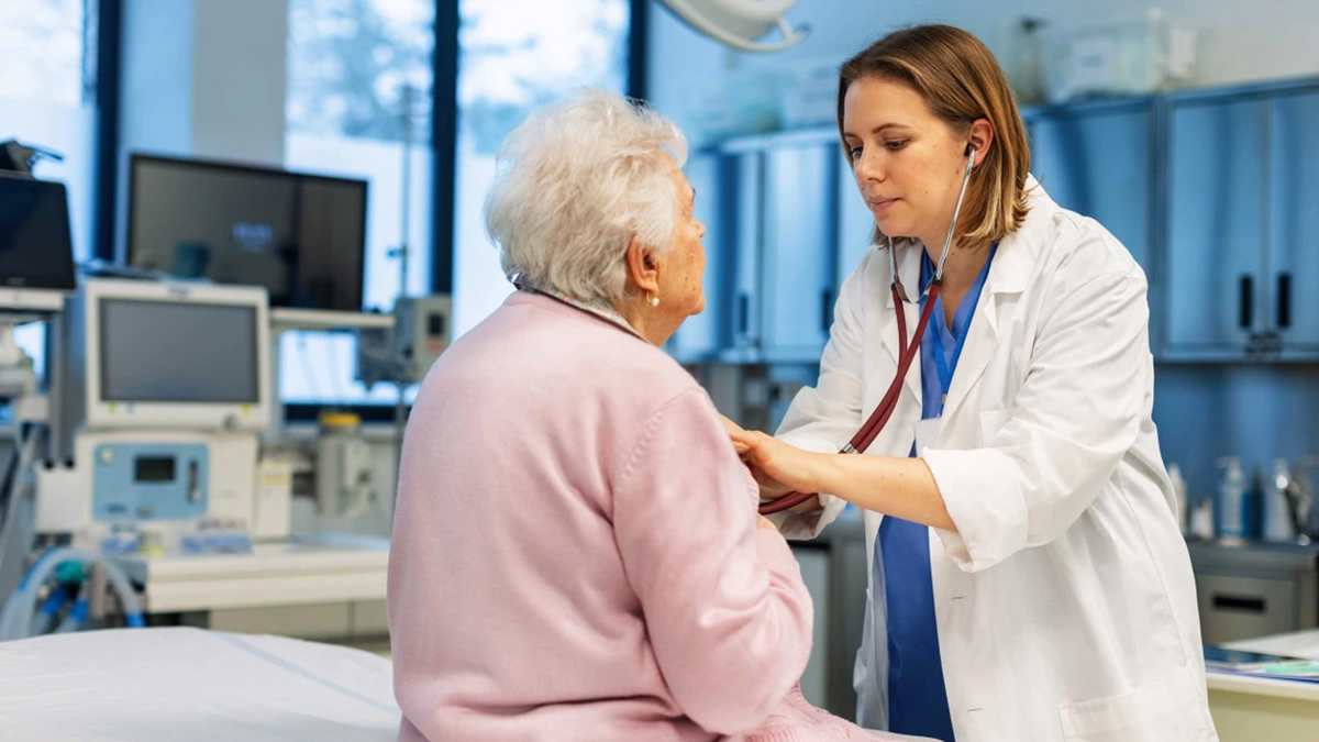 [A doctor examines a patient wearing a pink robe sitting on the edge of a bed]