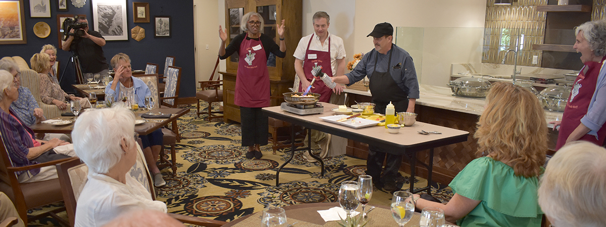 Two doctors wearing maroon aprons and the executive chef at the Hacienda at the River retirement community agog at the chef torching the top of ratatouille dish prepared for surrounding residents as a gluten-free option for controlled diets of those suffering from celiac disease.