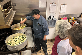 A man in black hat and gray chef’s jacket at a large stove stirs a pot of vegetables being prepared as ratatouille while an older woman and young man watch in the kitchen at the Hacienda at the River retirement community which served as locale for a cooking demonstration and fundraiser for the UArizona Division of Gastroenterology.