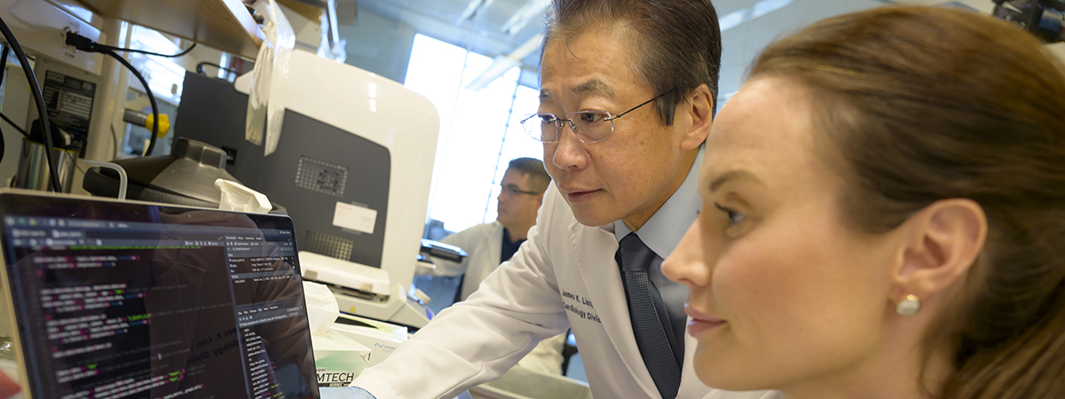 [A man in glasses and a lab coat looks over the shoulder of a woman in a lab coat and gloves as they examine information on a laptop.]