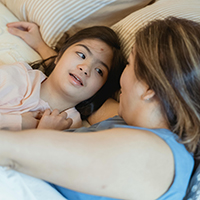 [A young girl looking at her mother while lying on the bed. (Source: Pexels.com/Nicola Barts)]