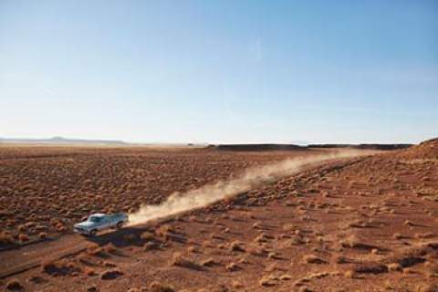 A pickup truck speeds down a dirt road in the desert on a hot day trailing dust behind it.