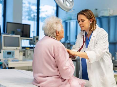 [A doctor examines a patient wearing a pink robe sitting on the edge of a bed]