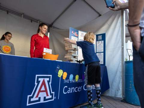 [Meet Yourself-1_skin cancer inst.jpg | Image Two students assist at the Skin Cancer event booth at Tucson Meet Yourself, a food festival held annually in October. A young boy plays a giant Jenga on the table while his father looks on and take a photo.]