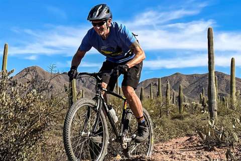 Older bicyclist mountain bikes through desert with Saguaro cactus in his wake.