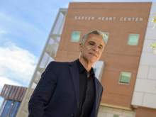 [A man in a dark suit coat and black shirt stands outside a building that has Sarver Heart Center signage.]