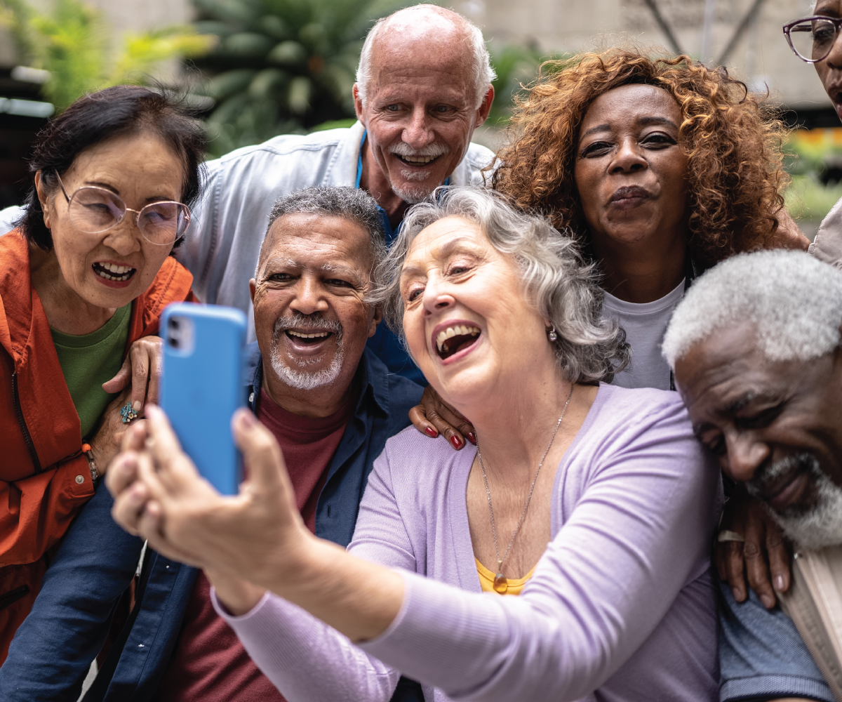 [Photo of a diverse group of older people gathering around a woman holding a cell phone for a selfie photograph.]