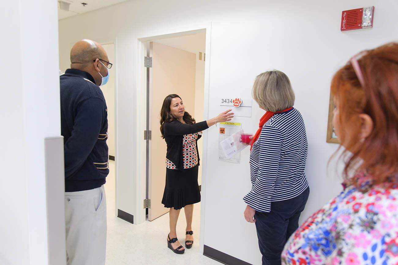 [A woman with dark hair points to signage outside an exam room during a tour with several people looking on.]