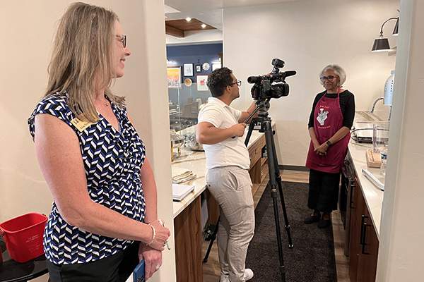 A blonde woman watches as a TV news camera person interviews a female physician wearing a maroon apron emblazoned with a "Cookin' Docs" logo about people with special dietary needs and how to add flavor to their meals while still eating healthy.