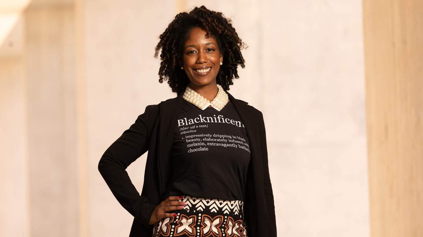 An African-American woman smiles with hand on hip wearing jacket and shirt that provides definition for "Blacknificent," standing in front of beige background.