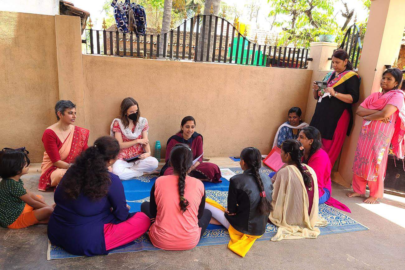 [Group of women wearing Indian saris sit in a circle having a discussion. There are two women standing off to the side, also wearing saris.]