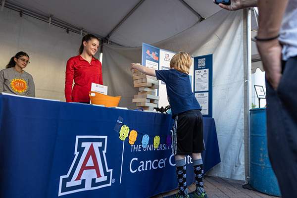 [Two students assist at the Skin Cancer event booth at Tucson Meet Yourself, a food festival held annually in October. A young boy plays a giant Jenga on the table while his father looks on and take a photo.]