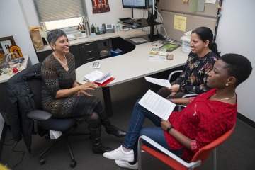 [South Asian professor sits at her desk talking with two of her mentees.]