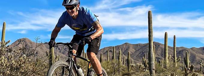 Older bicyclist mountain bikes through desert with Saguaro cactus in his wake 