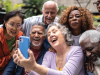 [Photo of a diverse group of older people gathering around a woman holding a cell phone for a selfie photograph.]